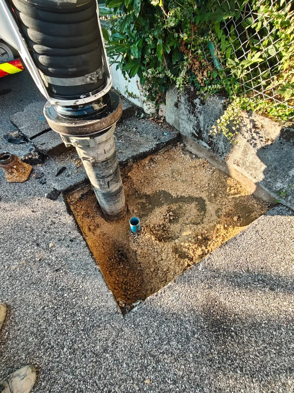Vue de dessus d’une fouille carrée dans l’enrobé : tuyau d’aspiration et conduite en PVC bleu émergeant du gravier